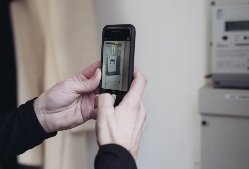 I billedet ser man to hænder, der holder en telefon og er i gang med at tage et billede af den elmåler, der hænger i baggrunden.