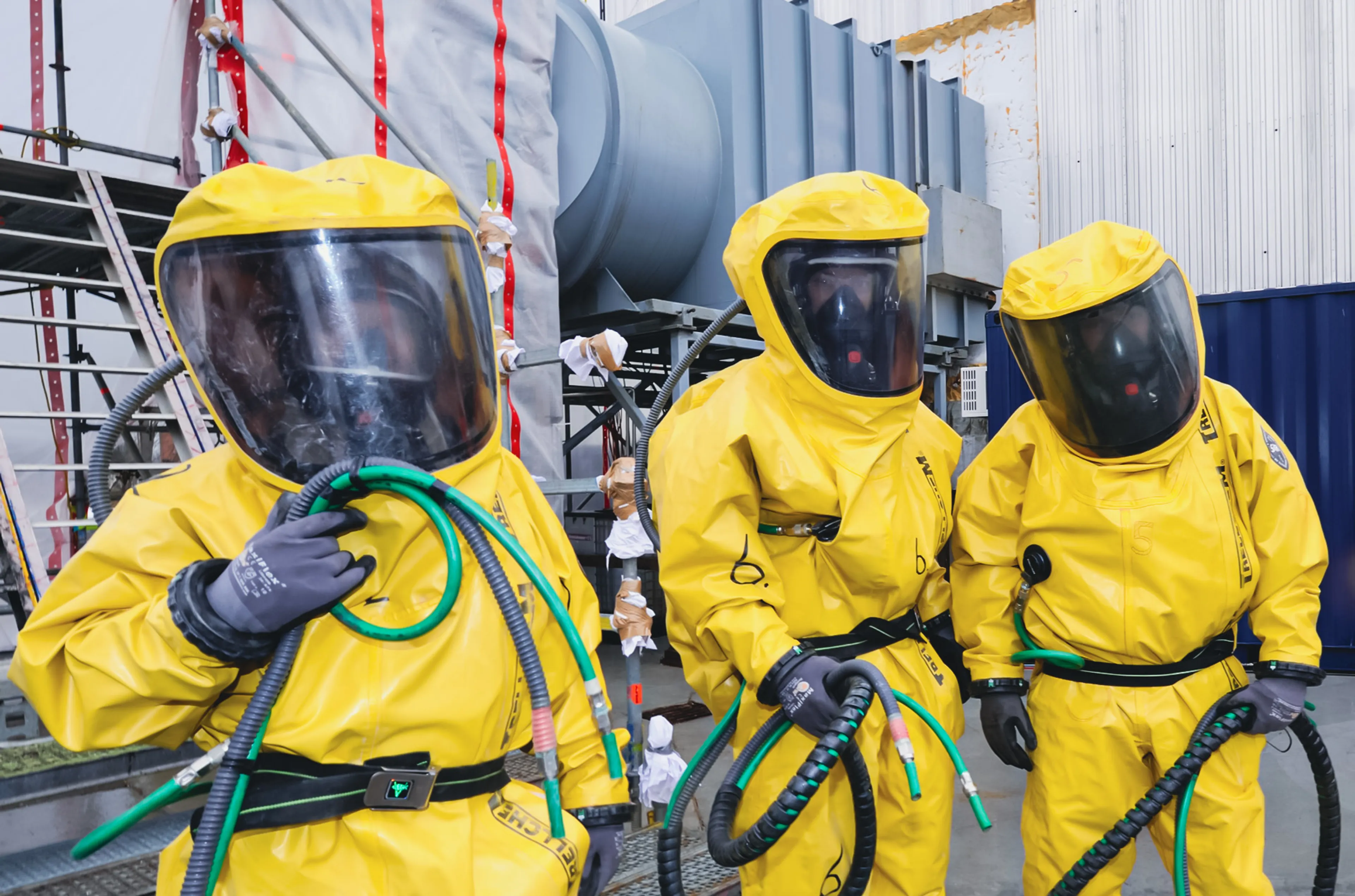Three technicians wearing yellow protective suits and full-face respirators prepare hoses and equipment during in-service inspection work inside an industrial ammonia storage facility. 