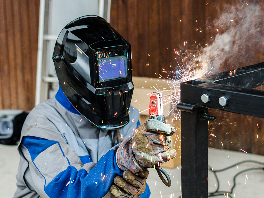 En svejser iført beskyttelsesudstyr udfører en svejsetest på en stålkomponent hvor der dannes gnister under aktiv svejsning.