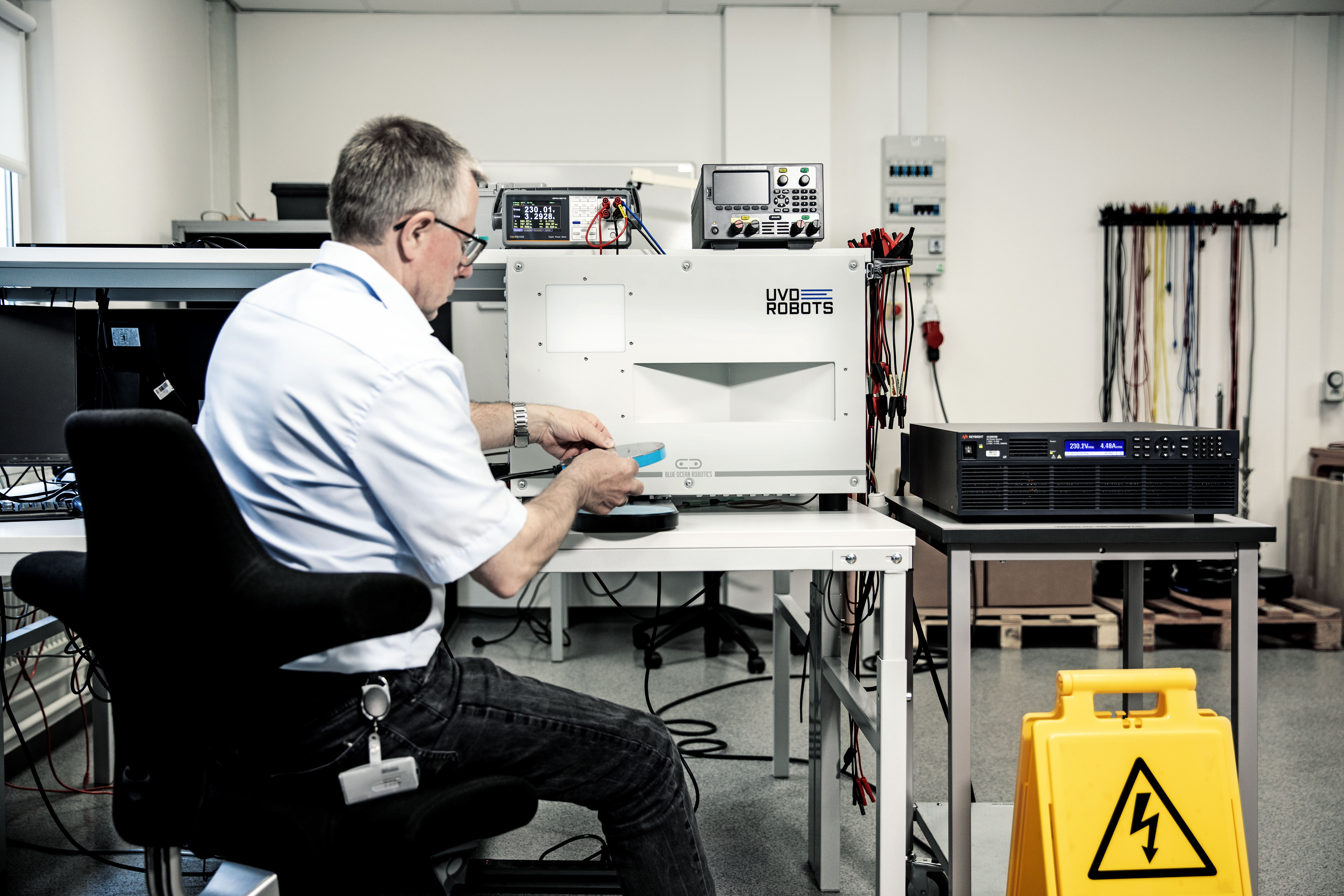 Technician performing electrical safety testing on an electronic product in a laboratory setup with measuring instruments and safety signage.