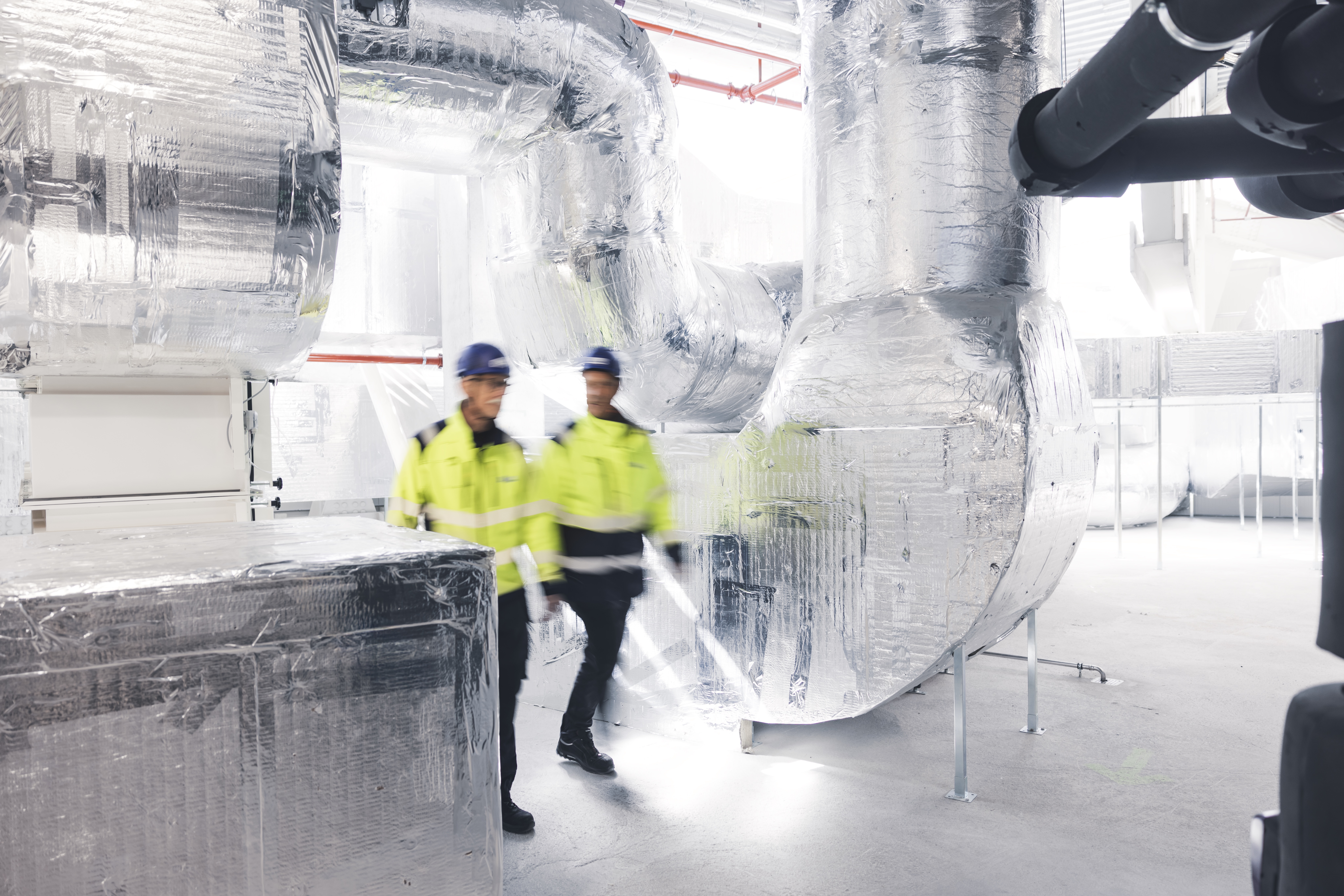 To teknikere i arbejdstøj går gennem et teknikrum med store isolerede ventilationskanaler og installationer, hvilket illustrerer syns- og skønsmænds arbejdsopgaver.