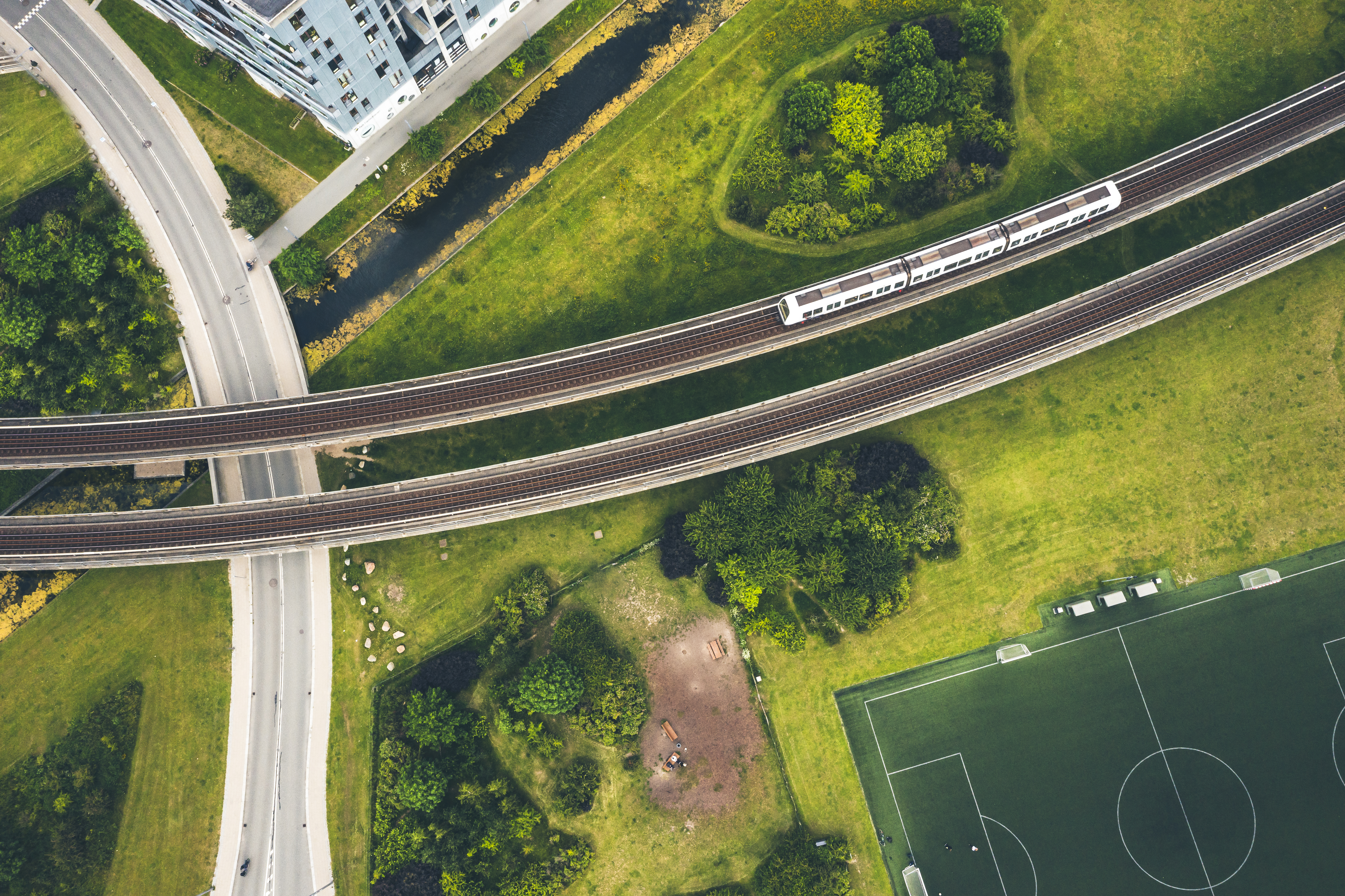 Aerial view of railway tracks with a train passing through a green urban area with roads, buildings and a football field, illustrating environmental context for noise measurement certification.