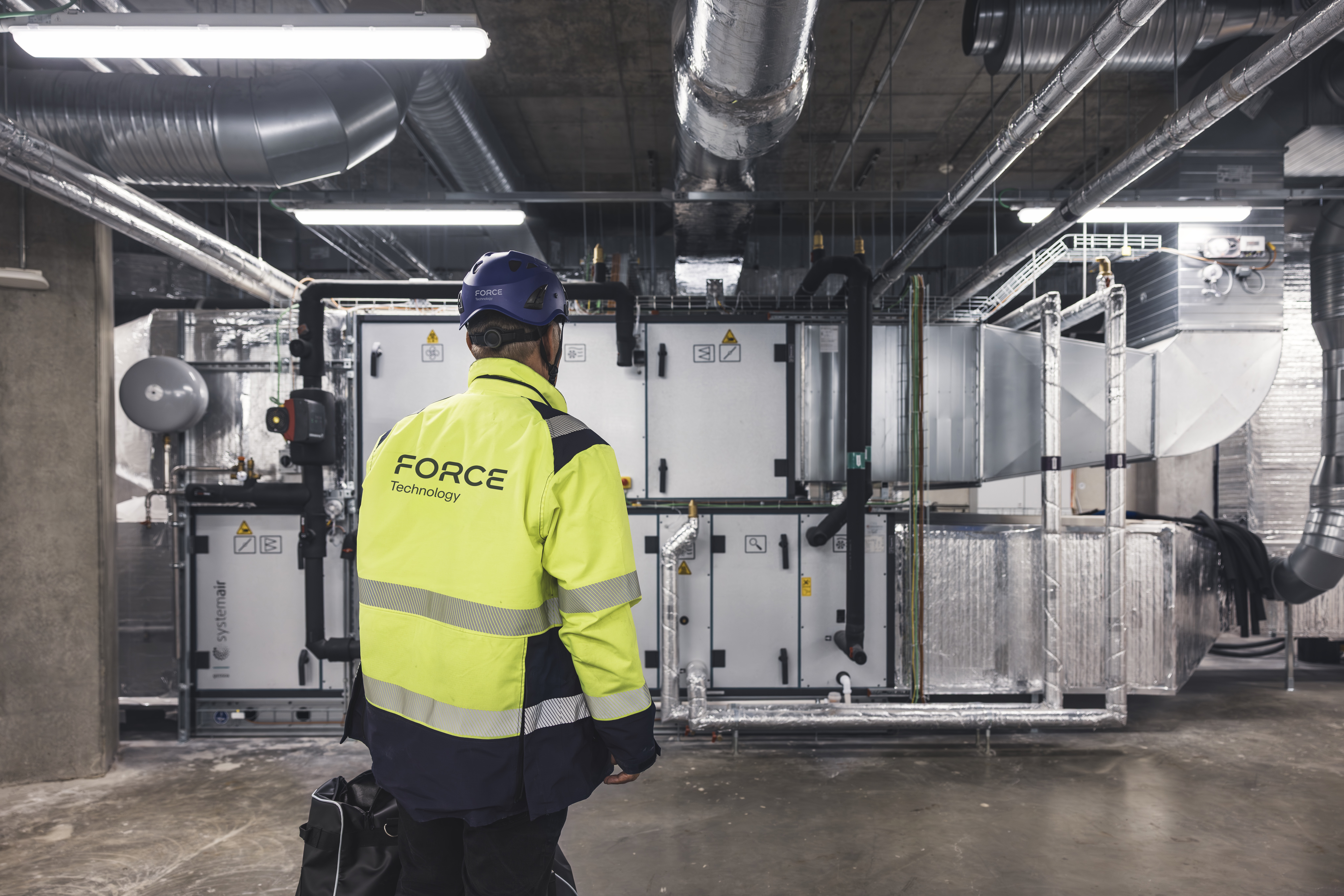 Technician in high-visibility jacket inspecting heating and ventilation equipment in a technical room with ductwork and energy installations.