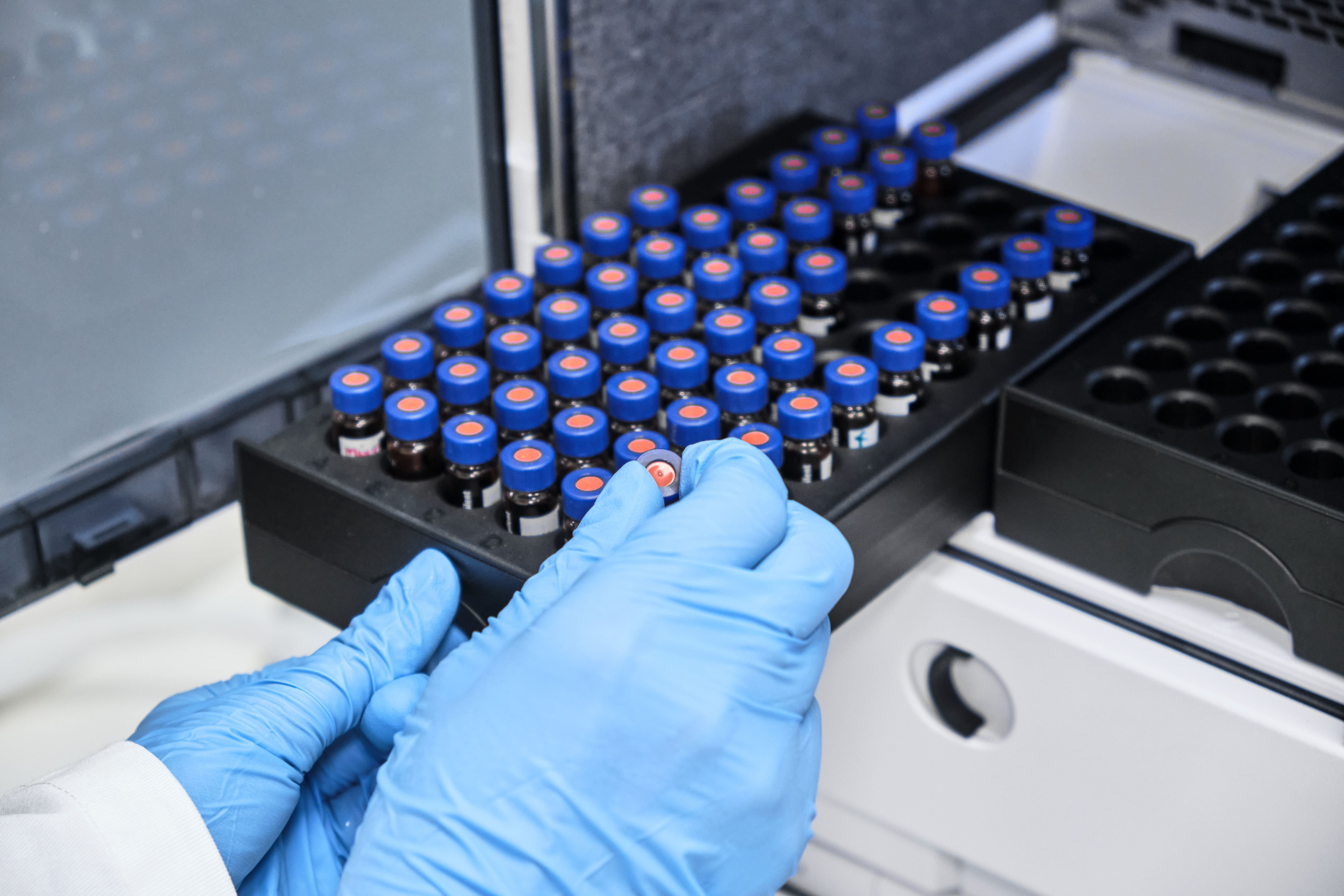 Specialist with blue-gloved hands places a sample vial into a laboratory tray before chemical analyses of consumer products.
