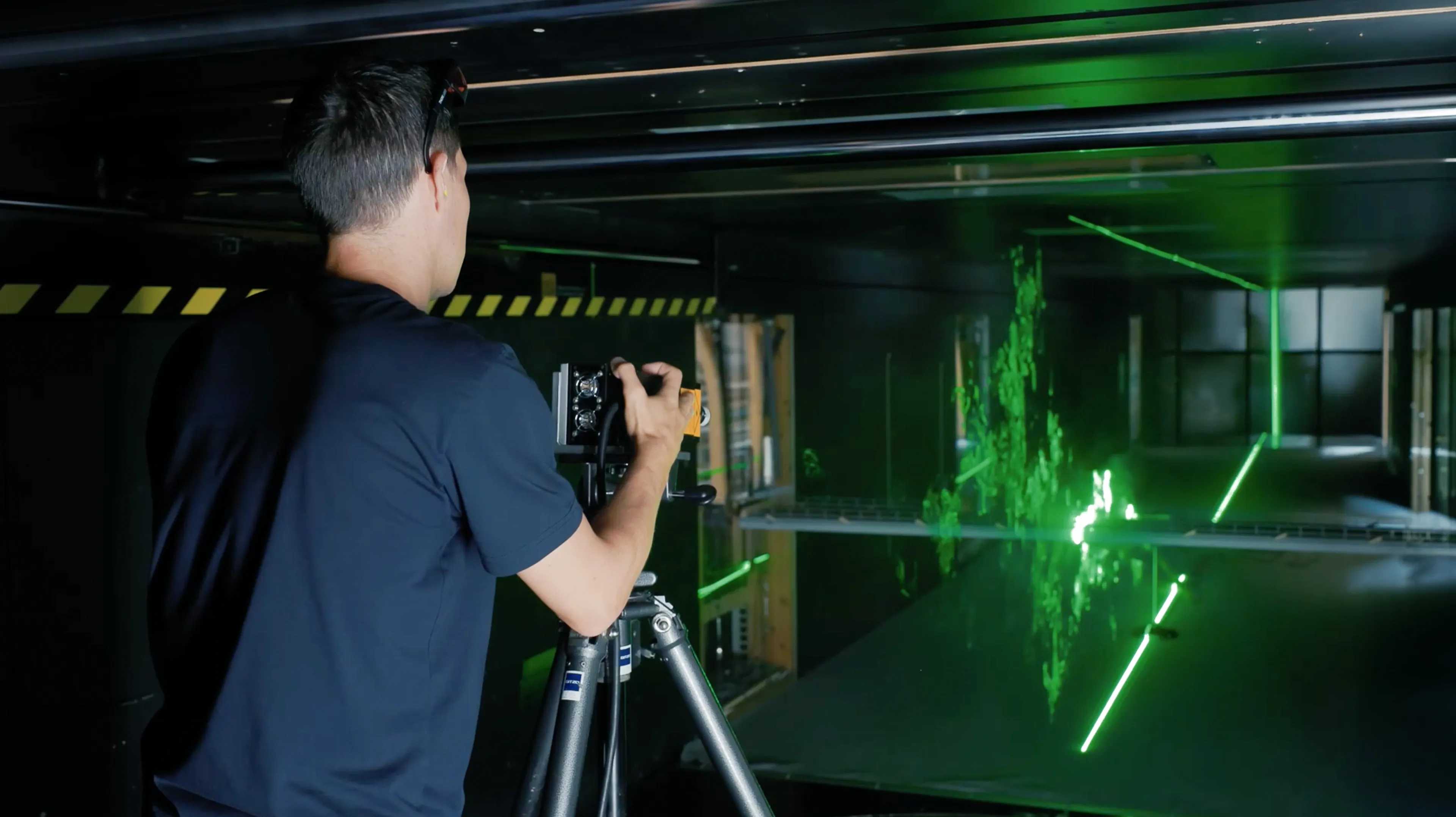 Technician using measurement equipment on a tripod inside a wind tunnel, observing green laser flow patterns around a bridge model during bridge aerodynamics testing.
