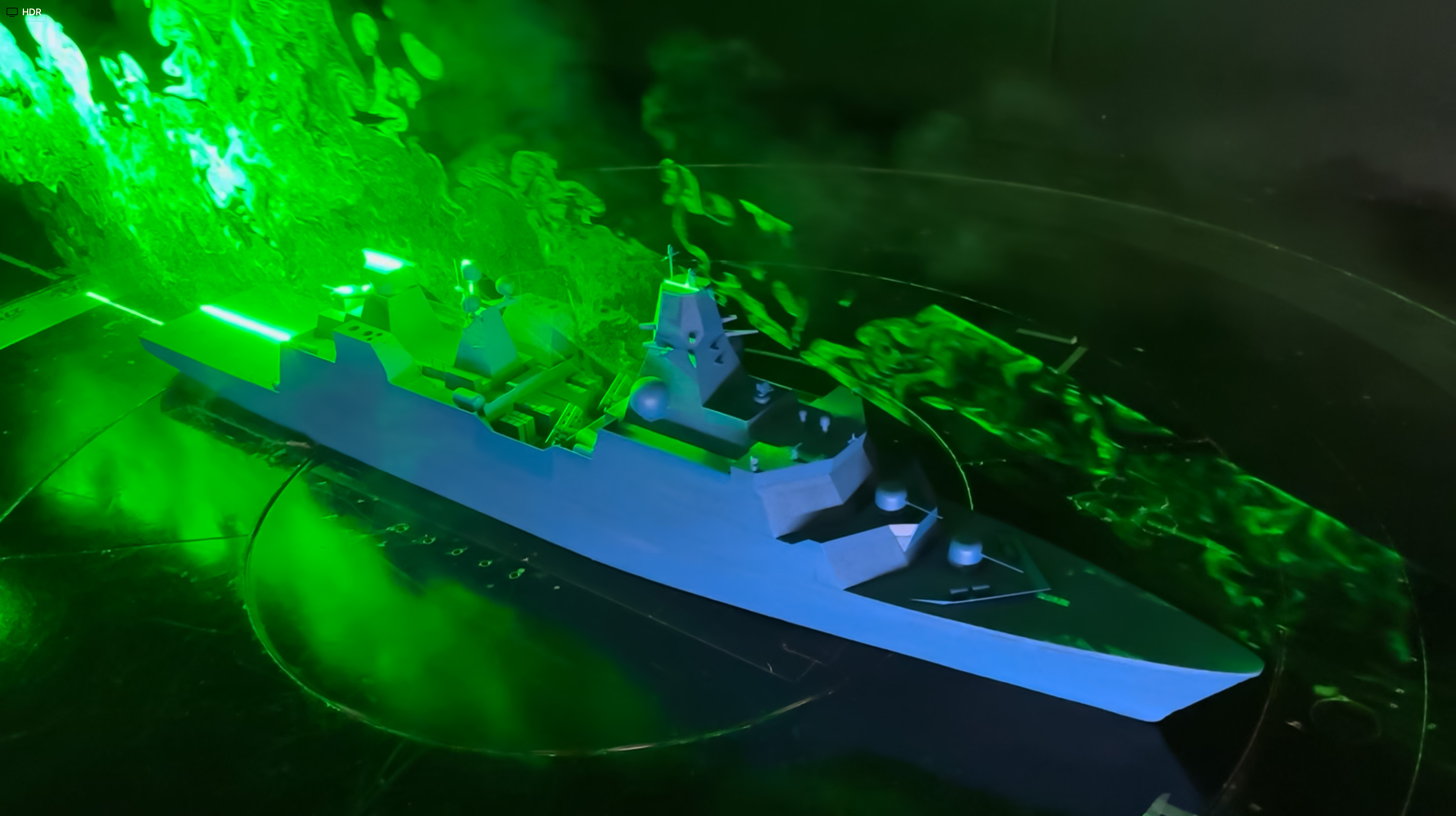 Scale model of a vessel in a wind tunnel with green flow visualisation showing airflow patterns around the ship structure during aerodynamic wind tunnel testing.