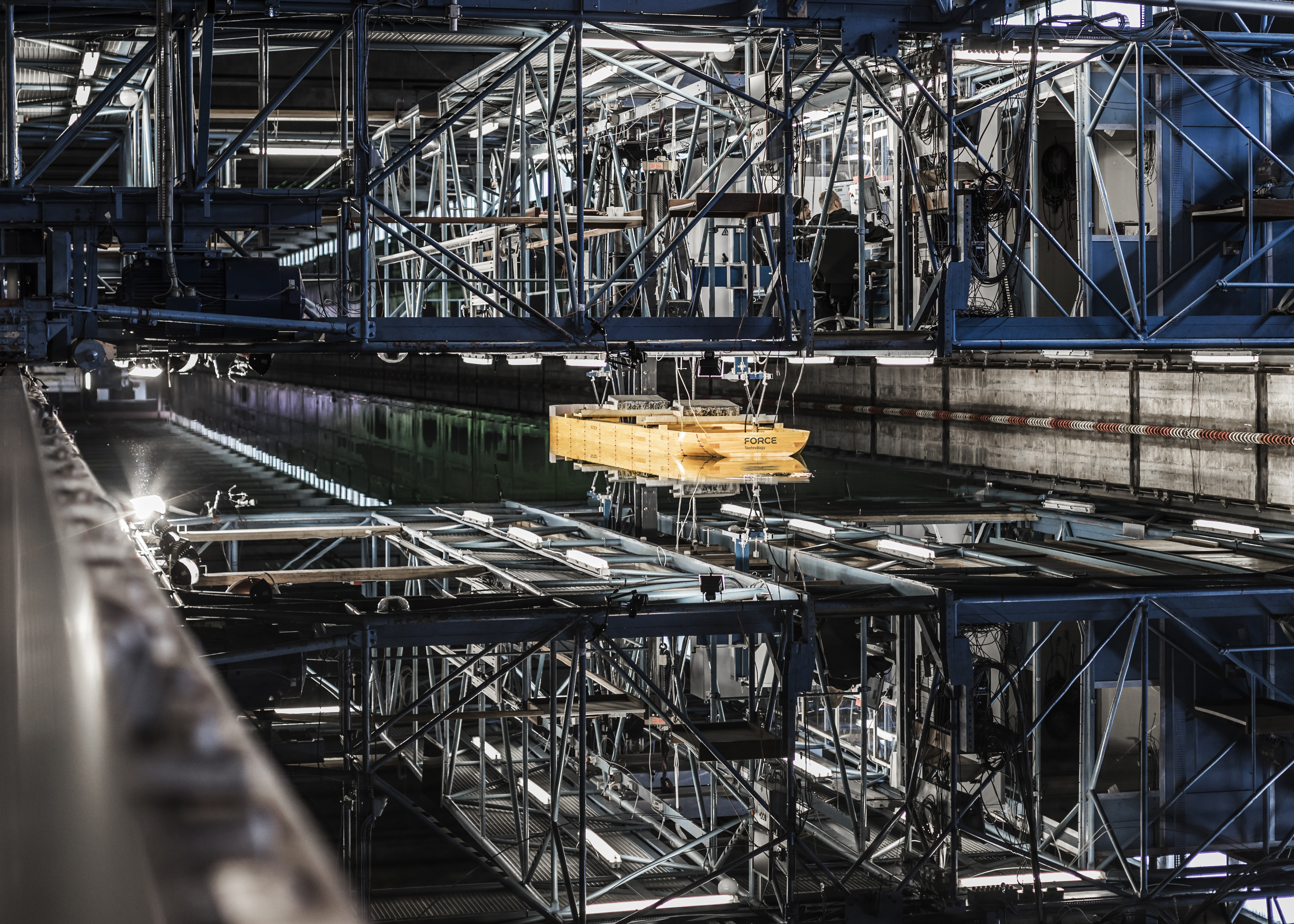 Indoor towing tank facility used for overtrawlability testing, showing a suspended model subsea structure being lowered into calm water beneath a steel test rig.