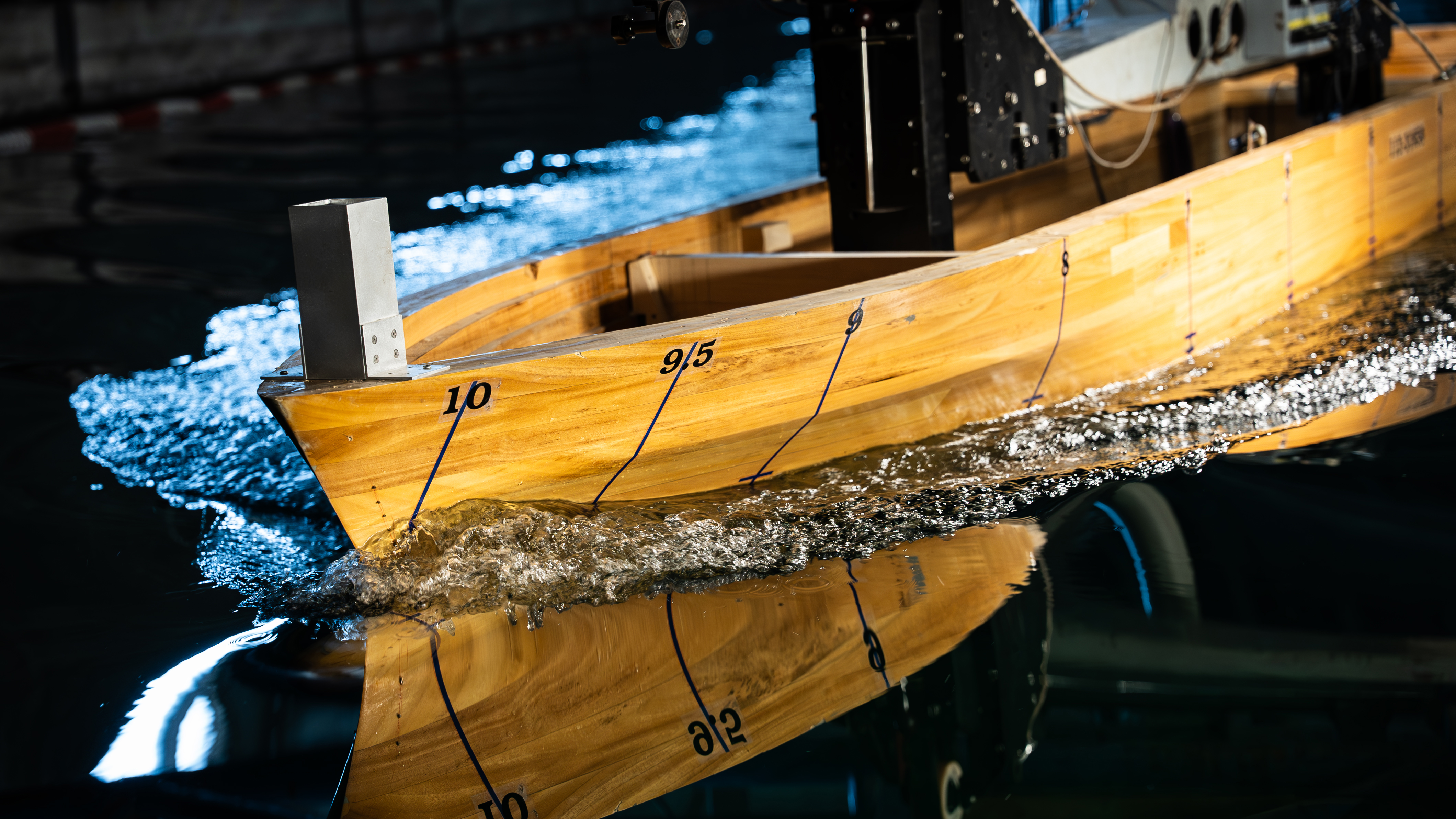 Scale model of a ship hull in a towing tank during manoeuvring evaluation, with markings along the hull and water flow visible along the bow.