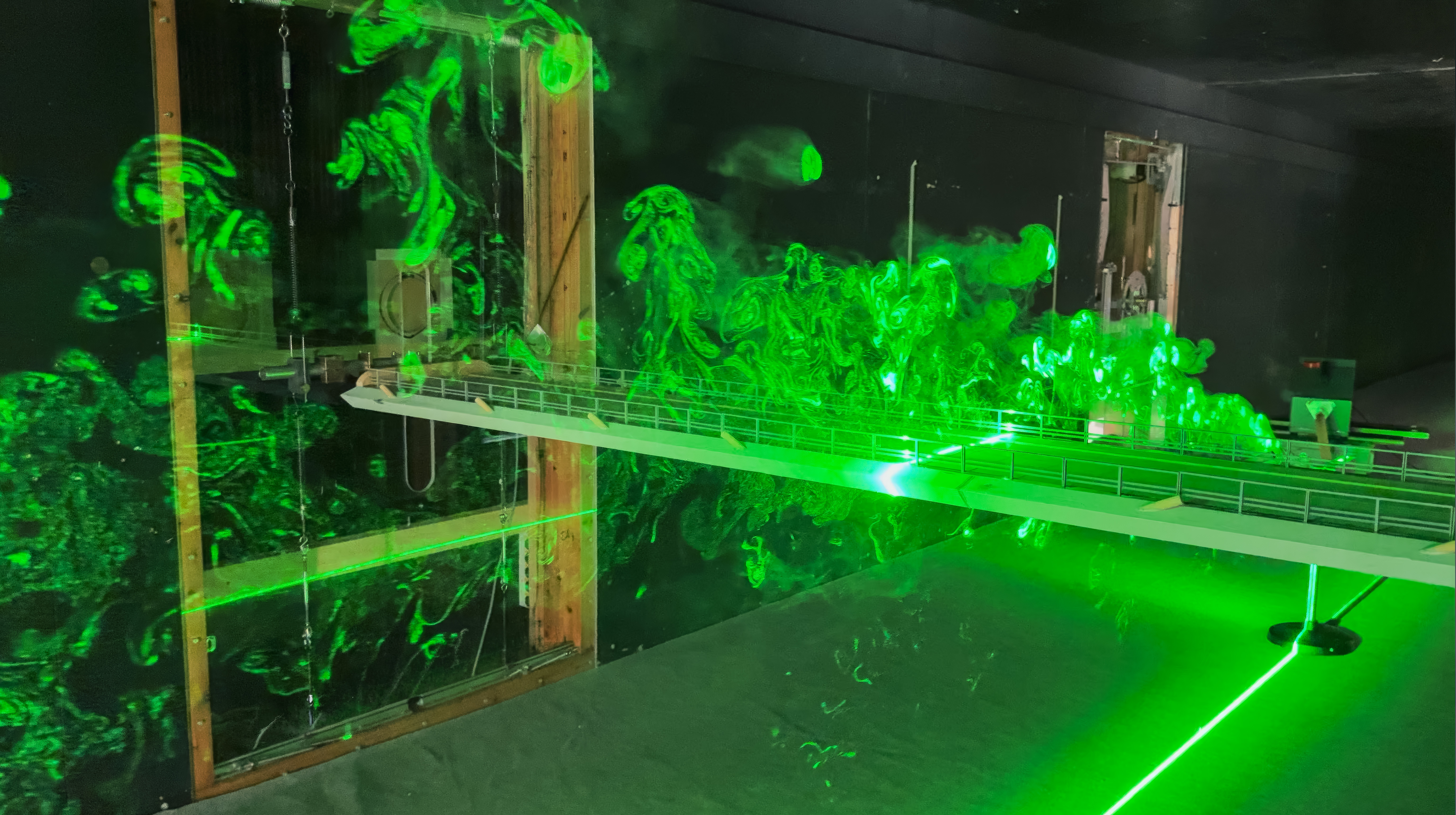 Vindtunnelopsætning med en skalamodel af en bro belyst med grønt laserlys, som viser luftstrømme og turbulens omkring konstruktionen.