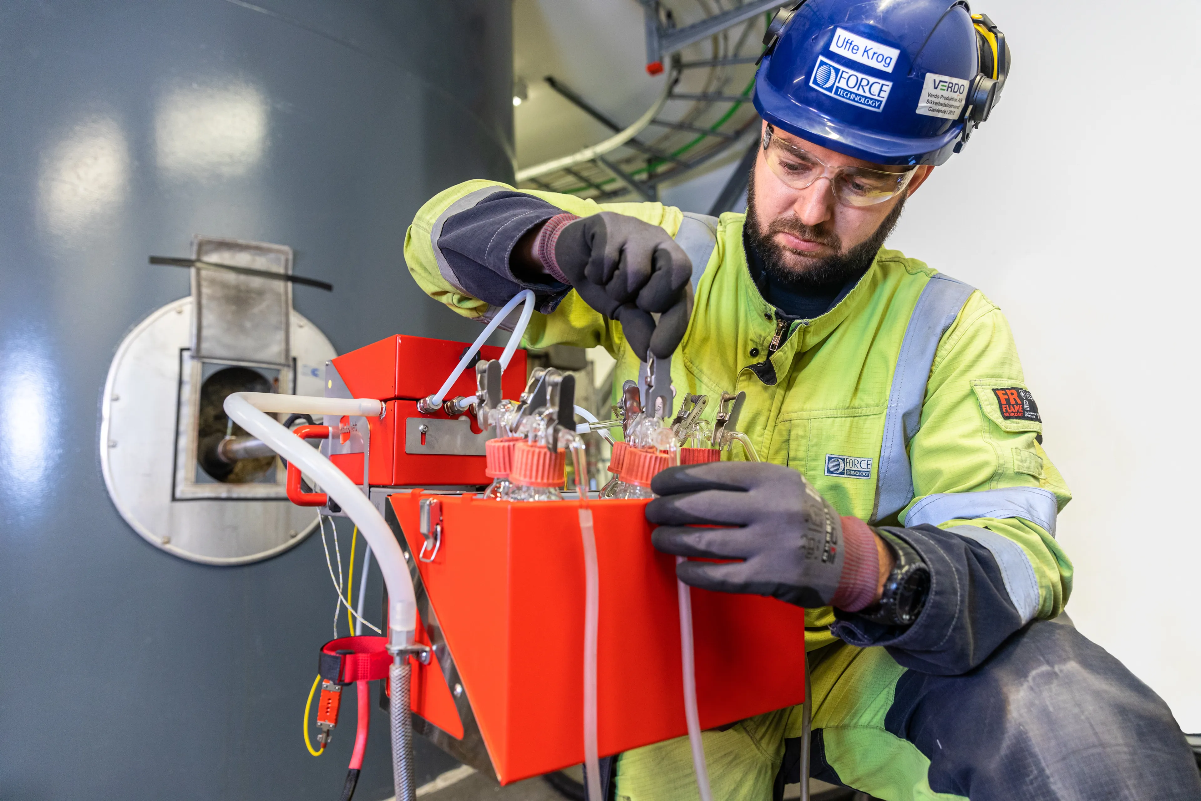 Technician wearing safety equipment collecting an air emission sample from a stack using measurement equipment with tubes and glass bottles. 