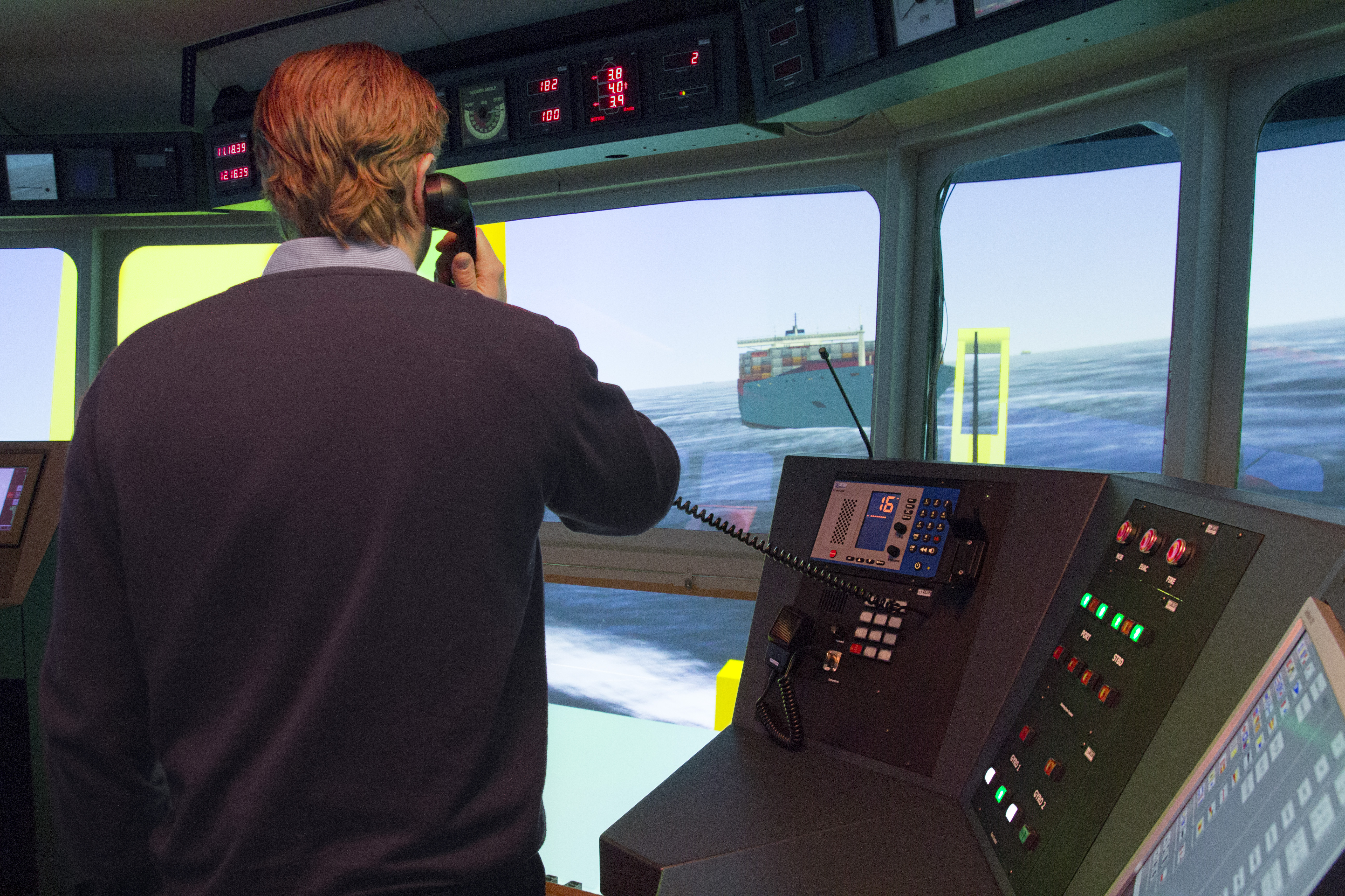 Person betjener udstyr i en skibsbro-simulator og ser et simuleret maritimt scenarie gennem broens vinduer.