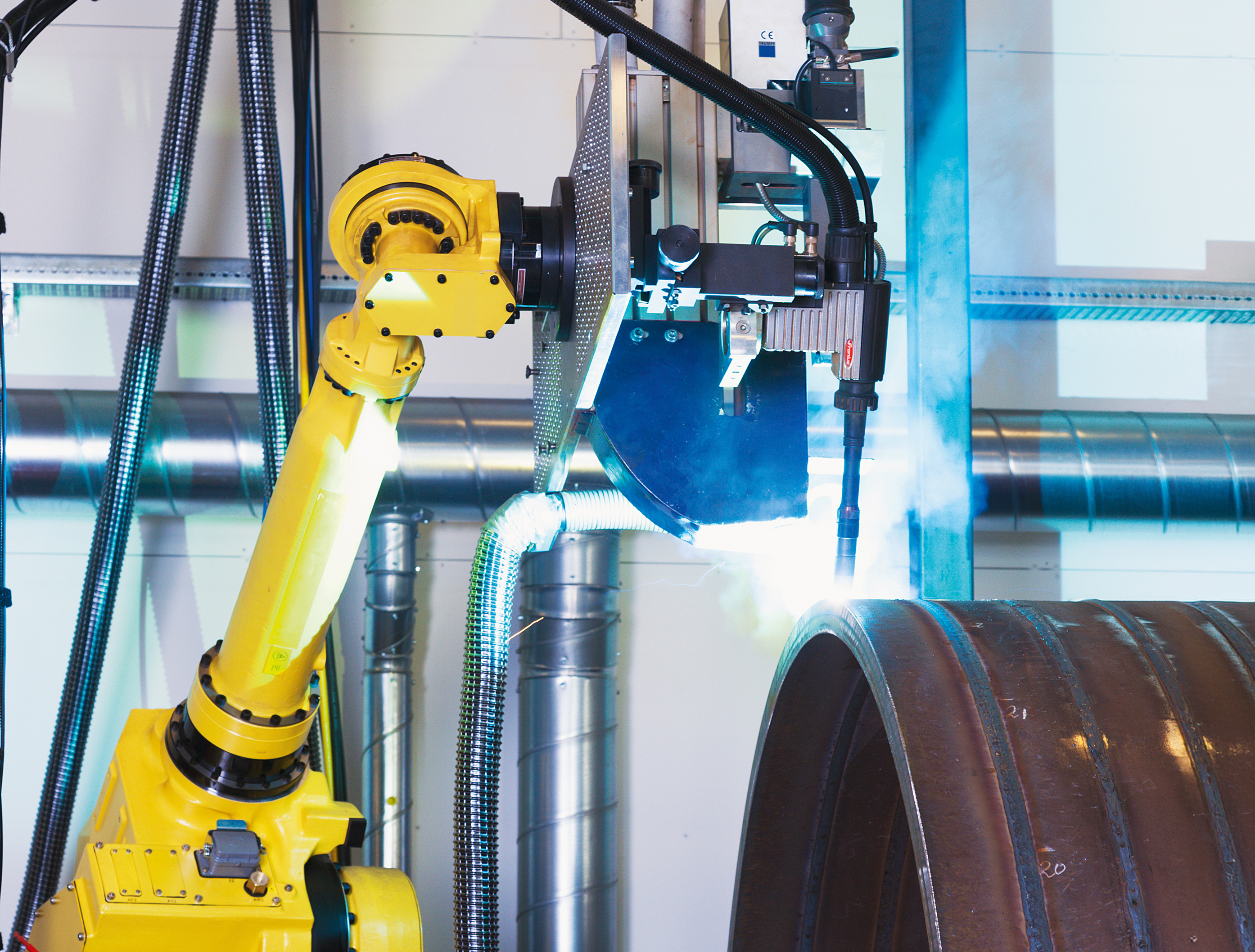 Yellow industrial robot performing laser welding on a large cylindrical metal component with visible welding light and extraction hoses.